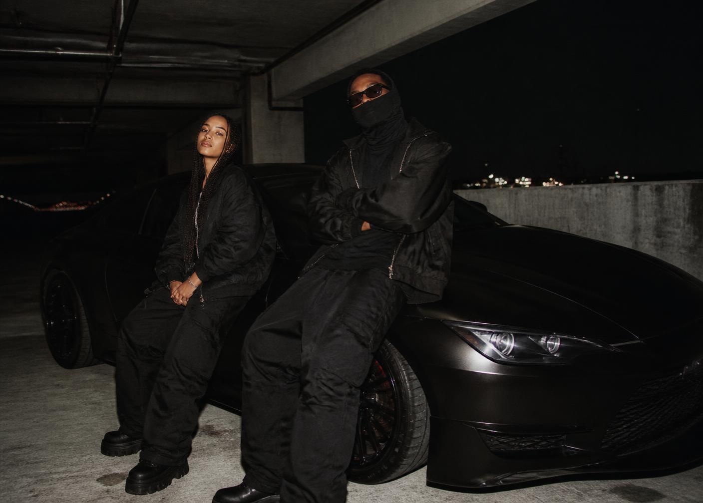 Two models in all black leaning on a matte black car in a parking garage